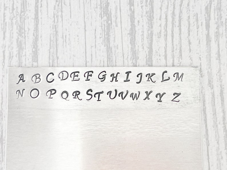 May include: A metal plate with the alphabet in cursive lettering. The letters are arranged in two rows, with the first row being A through M and the second row being N through Z.