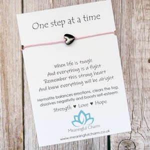 May include: A pink string bracelet with a black heart charm, attached to a white card. The card reads "One step at a time" and includes inspirational text about strength, love, and hope. The website address is also visible.