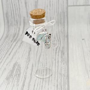 May include: A clear glass bottle with a cork stopper, a silver horse charm, and a tag that reads "POD Bear". The horse charm has a light blue color. The bottle is tied with white string.