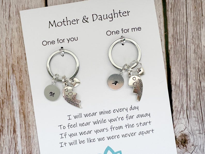 May include: Two silver-toned keychains attached to a white display card labeled Mother & Daughter. Each keychain features a circular ring, a small round charm with a letter, a heart-shaped charm, and a puzzle-piece heart charm engraved with Daughter.