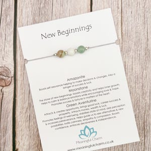 May include: A delicate string bracelet featuring three small, smooth, round beads in shades of brown, white, and green, mounted on a white display card labelled New Beginnings. The card includes text describing Amazonite, Moonstone, and Green Aventurine.