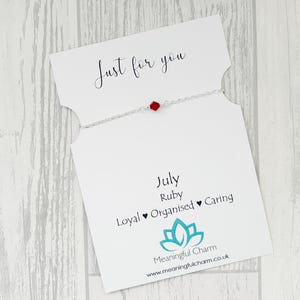 May include: A delicate silver chain bracelet featuring a single small red hexagonal bead, presented on a white card printed with the text Just for you, July, Ruby, Loyal, Organised, Caring, and the Meaningful Charm logo with its website address.