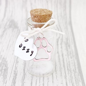 May include: A small glass jar with a cork stopper and a white ribbon tied around the neck. A silver paw print charm hangs from the ribbon, with a pink enamel fill. A white heart-shaped tag with the name "Ozzy" written on it is attached to the ribbon.