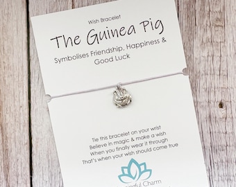 Puede incluir: Una pulsera de cordón blanco fino con un dije plateado en forma de cobaya, presentada en una tarjeta blanca con el texto The Guinea Pig, Symbolises Friendship, Happiness & Good Luck e instrucciones para pedir un deseo.
