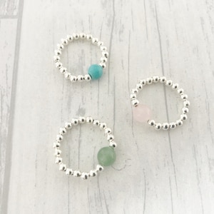 May include: Three elasticated rings made of small, polished silver-toned beads. Each ring features a single, larger round accent bead in a different colour: one turquoise blue, one pale pink, and one soft sage green, arranged in a circular formation.