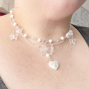 May include: A delicate silver-toned necklace featuring clear acrylic butterfly and bow charms, white pearl beads, and a heart-shaped pendant. The necklace is designed with multiple strands and is worn around the neck.