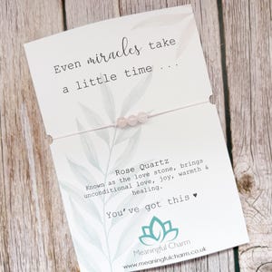 May include: A delicate bracelet featuring three small, pale pink rose quartz beads on a thin string, mounted on a white display card. The card displays the text "Even miracles take a little time...", "Rose Quartz", and "You've got this" with a logo.