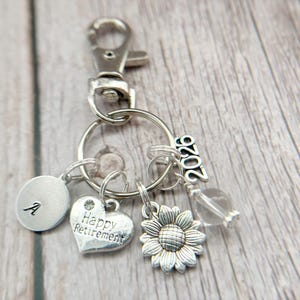 May include: Silver keychain with various charms. Charms include a heart with "Happy Retirement" text, a sunflower, a clear bead, an oval with the letter "A", and a "2020" charm. The keychain has a clasp for easy attachment.