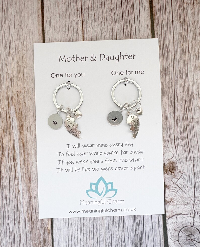May include: Two silver-toned keychains on a white display card labeled Mother & Daughter. Each keychain features a circular ring, a small round disc with a letter, a heart-shaped charm, and a puzzle piece charm engraved with the word Mother or Daughter.