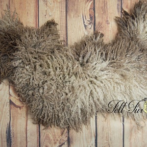May include: A sheepskin rug with a natural, curly texture in shades of brown and cream. The rug is laid on a wooden plank background, showcasing its organic shape and soft appearance. The logo "Felt Fur" is visible in the bottom right corner.