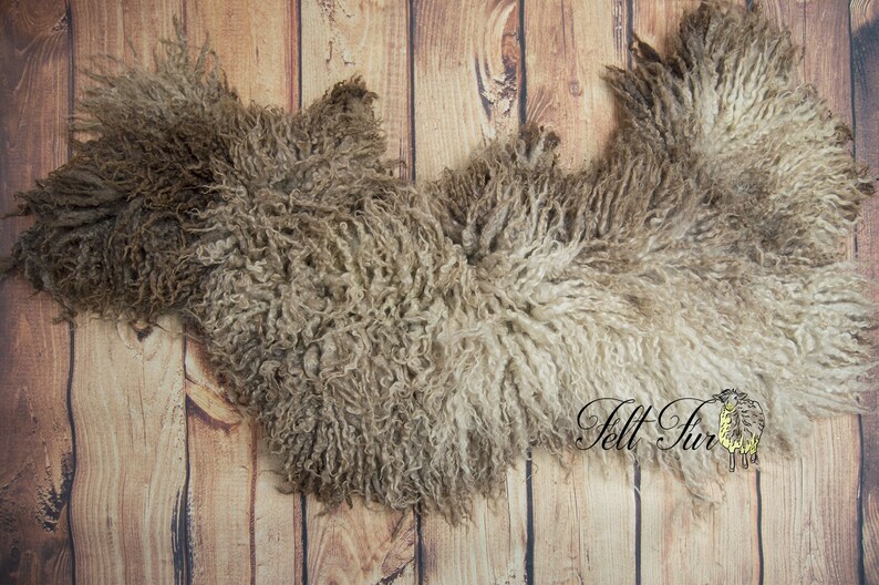 May include: A sheepskin rug with a natural, curly texture in shades of brown and cream. The rug is laid on a wooden plank background, showcasing its organic shape and soft appearance. The logo "Felt Fur" is visible in the bottom right corner.
