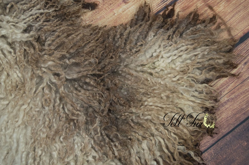 May include: Close-up of a sheepskin pelt with a mix of light and dark brown curly wool. The texture is dense and wavy, with variations in color and depth. The background is a wooden surface.
