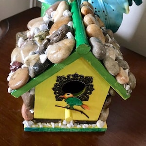 May include: A yellow birdhouse with a green roof and a bird on a branch. The birdhouse is decorated with small stones and a metal design.