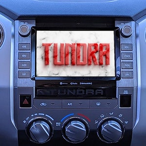 Op de afbeelding: Een close-up van een dashboard van een auto met een scherm waarop het woord "Tundra" in rode letters wordt weergegeven. Het scherm wordt omringd door knoppen en bedieningselementen, waaronder een alarmknop, een klimaatregelpaneel en een volumeknop.