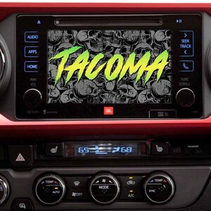 May include: A black car dashboard with a digital display screen showing the word "TACOMA" in green and yellow letters on a black and white skull background. The screen is surrounded by buttons labeled "AUDIO", "APPS", "HOME", "PWR VOL", "SEEK TRACK", "TUNE SCROLL", and "JBL".