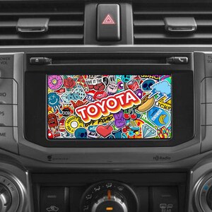 Könnte beinhalten: Ein Armaturenbrett eines Autos mit einem Touchscreen-Display, das ein farbenfrohes Bild von Aufklebern mit dem Wort "TOYOTA" in der Mitte zeigt. Das Display ist von Knöpfen für Audio, Apps, Home, Suchen, Track und Tune Scroll umgeben.