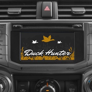 Puede incluir: Un tablero de instrumentos de un coche con una pantalla que muestra el texto "Duck Hunter" en una fuente estilizada. La pantalla también muestra un gráfico de un pato volando sobre un campo de hierba alta.