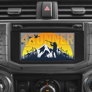 Può includere: Un sistema stereo per auto con uno schermo che mostra una scena di caccia con la scritta "4Runner". La scena presenta un cacciatore, un cane, montagne e uccelli in volo, sullo sfondo di un tramonto giallo e arancione.