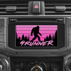 Może przedstawiać: Czarno-różowa deska rozdzielcza samochodu z ekranem wyświetlającym sylwetkę Bigfoota idącego przed zachodem słońca z napisem "4RUNNER".