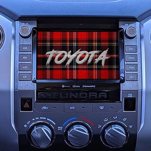 Puede incluir: Panel de instrumentos de coche con una pantalla que muestra la palabra "TOYOTA" en una fuente estilizada sobre un patrón de cuadros rojo y negro. El salpicadero tiene botones y mandos.