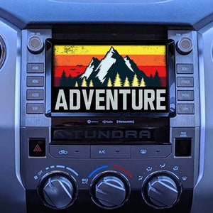 以下が含まれることがあります： 山と「ADVENTURE」の言葉がレトロなスタイルで描かれた画面が表示されている車のダッシュボード。画面はボタンとコントロールで囲まれています。