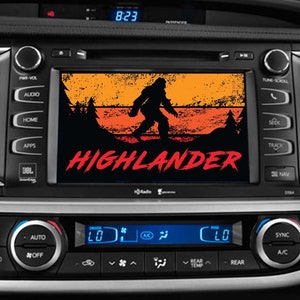May include: A car's dashboard with a screen displaying a retro-style graphic of a Bigfoot with the text "HIGHLANDER" in red. The screen is surrounded by buttons and controls.