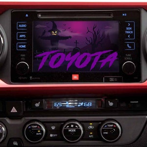 May include: A car dashboard with a screen displaying a Halloween-themed image of a scarecrow and a graveyard. The screen also displays the word "TOYOTA" in purple letters. The dashboard has buttons for audio, apps, home, power, volume, temperature, mode, and air conditioning.