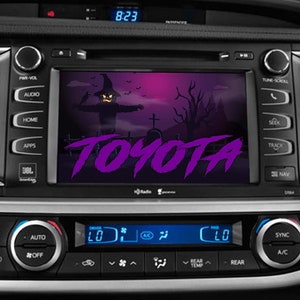 May include: A car's dashboard with a screen displaying a Halloween-themed image of a scarecrow with a purple "TOYOTA" logo. The screen is surrounded by buttons and controls for the car's audio system and climate control.