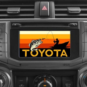Op de afbeelding: Een autoscherm toont een visscène met een springende vis en een visser in een boot tegen een zonsondergang. Het woord "TOYOTA" staat in gele blokletters eronder. De stereo zit in een grijs dashboard.