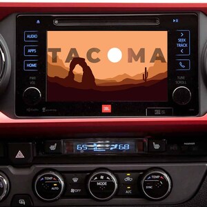 Peut inclure: Gros plan sur le tableau de bord d'une voiture avec un écran tactile affichant le mot "TACOMA" dans une police stylisée. L'écran affiche également une scène de coucher de soleil avec une arche de montagne en arrière-plan. Le tableau de bord comporte des boutons pour l'audio, les applications, la maison, l'alimentation, le volume, la température et d'autres commandes.