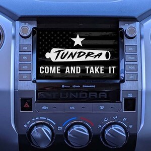 以下が含まれることがあります： 車のダッシュボードの白黒画像で、画面には大砲のスタイリッシュな画像と「Tundra Come and Take It」というテキストが表示されています。