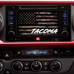 May include: A black and white American flag with a camouflage pattern overlay, with the word "TACOMA" in white text on a black background, displayed on a car's dashboard screen.