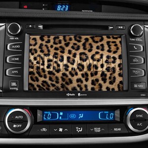 May include: A car dashboard with a screen displaying a leopard print background with the word "HIGHLANDER" in white text. The screen is surrounded by buttons and controls for the car's audio system, climate control, and navigation.