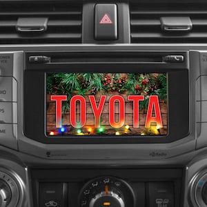 May include: A car's dashboard with a screen displaying the word "TOYOTA" in red letters. The screen shows a festive holiday scene with pine branches, berries, and string lights. The dashboard has various buttons and knobs.