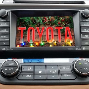 May include: A car dashboard with a screen displaying the word "TOYOTA" in red letters on a wooden background with Christmas lights. The screen is surrounded by buttons for audio, apps, home, and other functions. The dashboard also has climate controls with a digital display showing 24.0 degrees.