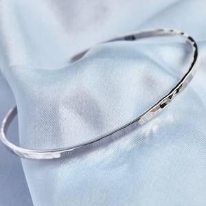 Op de afbeelding: Een zilverkleurige, gehamerde armband. Het open manchetontwerp wordt gepresenteerd op een lichtblauwe satijnen stof. De armband heeft een eenvoudige, elegante stijl, geschikt voor dagelijks gebruik.