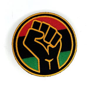 May include: Embroidered patch featuring a black fist raised in solidarity, outlined in gold against a circular background. The background is divided into red, black, and green horizontal stripes, with a gold border.