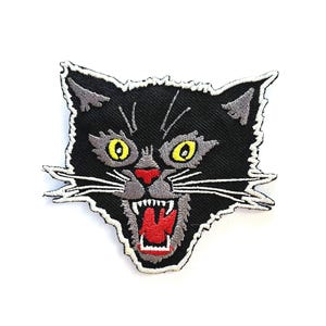 May include: Embroidered patch featuring a black cat head with yellow eyes, a red nose, and an open mouth displaying teeth. The cat has gray accents and white whiskers, with a white border.