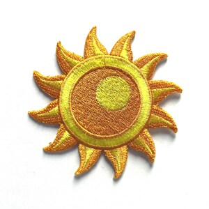 May include: Embroidered patch featuring a stylized sun design. The sun has a golden yellow outer ring with pointed rays and a central orange circle with a lighter yellow center. The patch is on a white background.