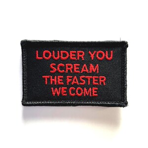 May include: Black rectangular patch with red embroidered text that reads "LOUDER YOU SCREAM THE FASTER WE COME". The patch has a black border and is likely designed to be sewn or ironed onto clothing or accessories.