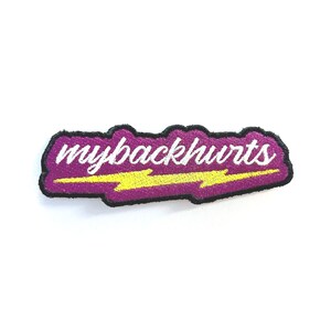May include: Embroidered patch with the text "mybackhurts" in white script, outlined in black, on a purple background. A yellow lightning bolt graphic is beneath the text. The patch is rectangular with rounded edges.