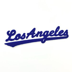 May include: A blue and white patch featuring the script text "Los Angeles". The text is in a cursive font with a white outline and a blue felt-like texture. The patch has a white background.