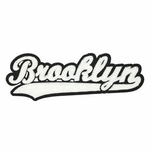 May include: A white and black Brooklyn patch with a script font. The word "Brooklyn" is in a cursive style, with a white, textured center and a black border. The patch has a curved, elongated shape.