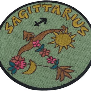 May include: Round patch with the word "SAGITTARIUS" in yellow letters. The patch features a brown bow and arrow, a yellow sun, a crescent moon, and pink flowers on a light green background, with a black border.
