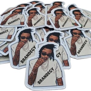 May include: A pile of white patches with a cartoon illustration of a person wearing sunglasses and a white shirt. The patches have a white border and the word "BRANDEGY" is printed on the shirt.