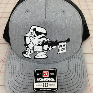 May include: A gray and black trucker hat with a white cartoon image of a Stormtrooper holding a blaster and a flag that says "PEW PEW". The hat has a black mesh back and a gray front with the text "EST. 1970 RICHARDSON. ORIGINAL 112 TRUCKER ADJUSTABLE" on a black tag.