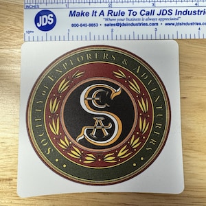 May include: A square sticker with a circular emblem featuring the text "SOCIETY OF EXPLORERS & ADVENTURERS." The emblem is in a maroon, gold, and white color scheme. A ruler is placed above the sticker, measuring approximately 2 inches.