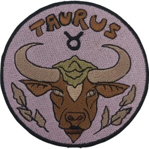 May include: Embroidered patch featuring the Taurus zodiac symbol. The design includes a brown bull's head with large horns, surrounded by a light purple circle with the word "TAURUS" in brown lettering. A black border frames the patch.
