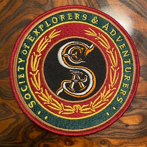 May include: A round, embroidered patch with a red, green, and gold border. The center features a black background with a white and orange stylized letter "S" and the text "Society of Explorers & Adventurers".