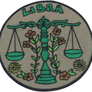 May include: Embroidered patch featuring the Libra zodiac symbol. The design includes a green scale, floral accents, and the word "LIBRA" in green lettering on a tan background. The patch has a black border.